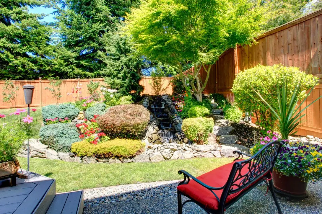 A beautifully landscaped backyard featuring a small rock waterfall cascading into a pebble-lined stream. The garden is lush with various shrubs, plants, and colorful flowers. A red-cushioned bench faces the waterfall, positioned on a gravel path beside green grass.