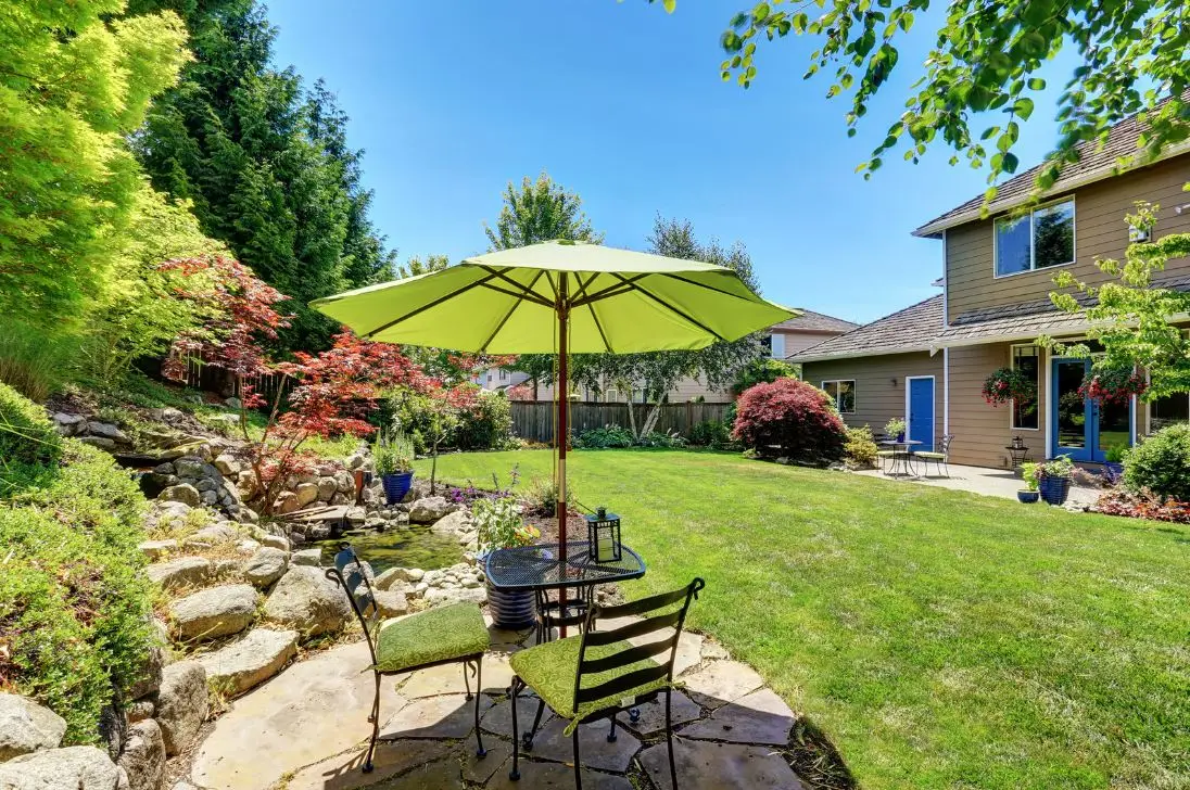A tranquil backyard scene featuring a patio with a round table and two green cushioned chairs under a bright green umbrella. The patio is surrounded by lush greenery, stone steps, and various plants. A beige house with large windows is visible in the background.